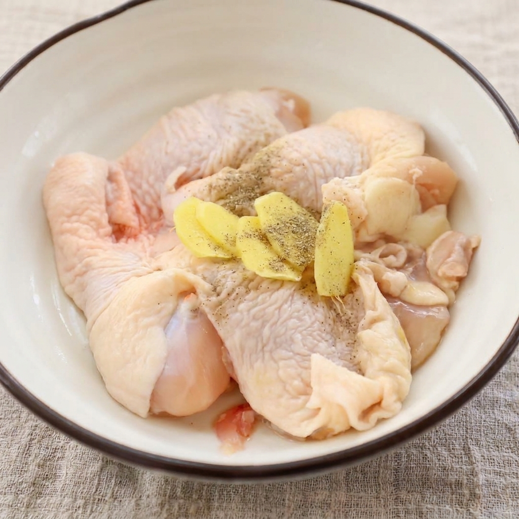 Rohe Hähnchenstücke in einer Schüssel, bestreut mit frischem Ingwer und Pfeffer.