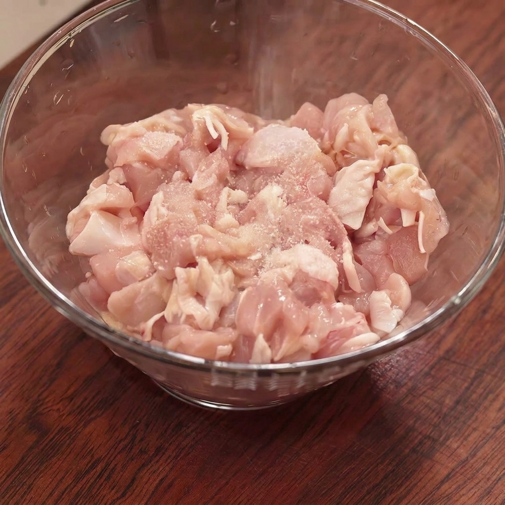 Rohes, gehacktes Hähnchen in einer Glasschüssel, bestreut mit Salz, Speisestärke und Gewürzen vor dem Vermengen.