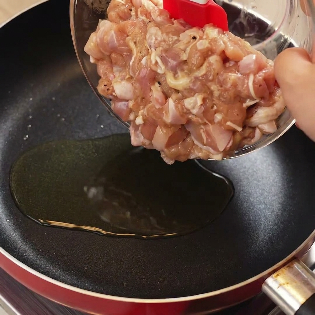 Mit einem roten Pfannenwender werden marinierte, rohe Hähnchenstücke aus einer Schüssel in eine heiße Pfanne mit Öl gegeben.