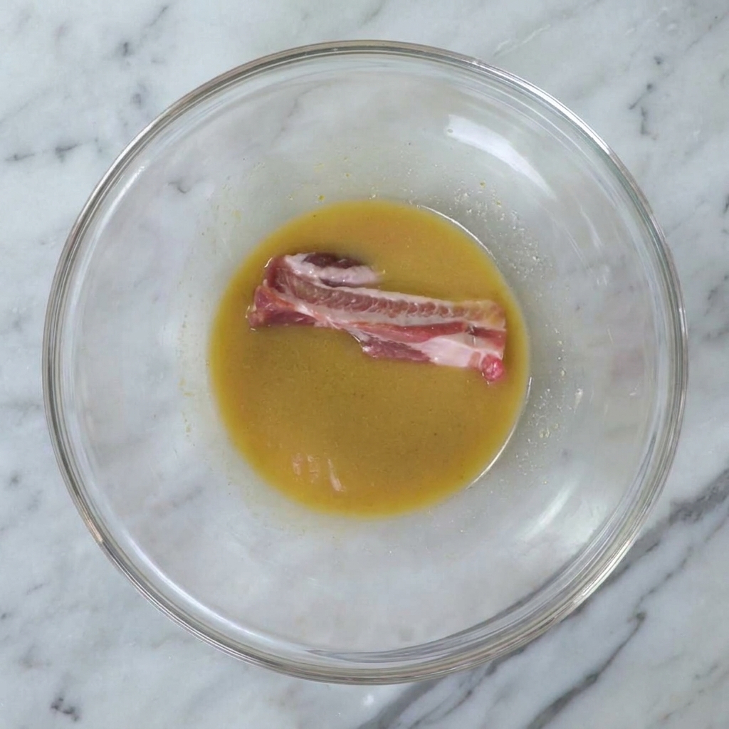 Ein rohes Schweinerippchen wird in die goldene Miso-Marinade in einer großen Glasschüssel getaucht.