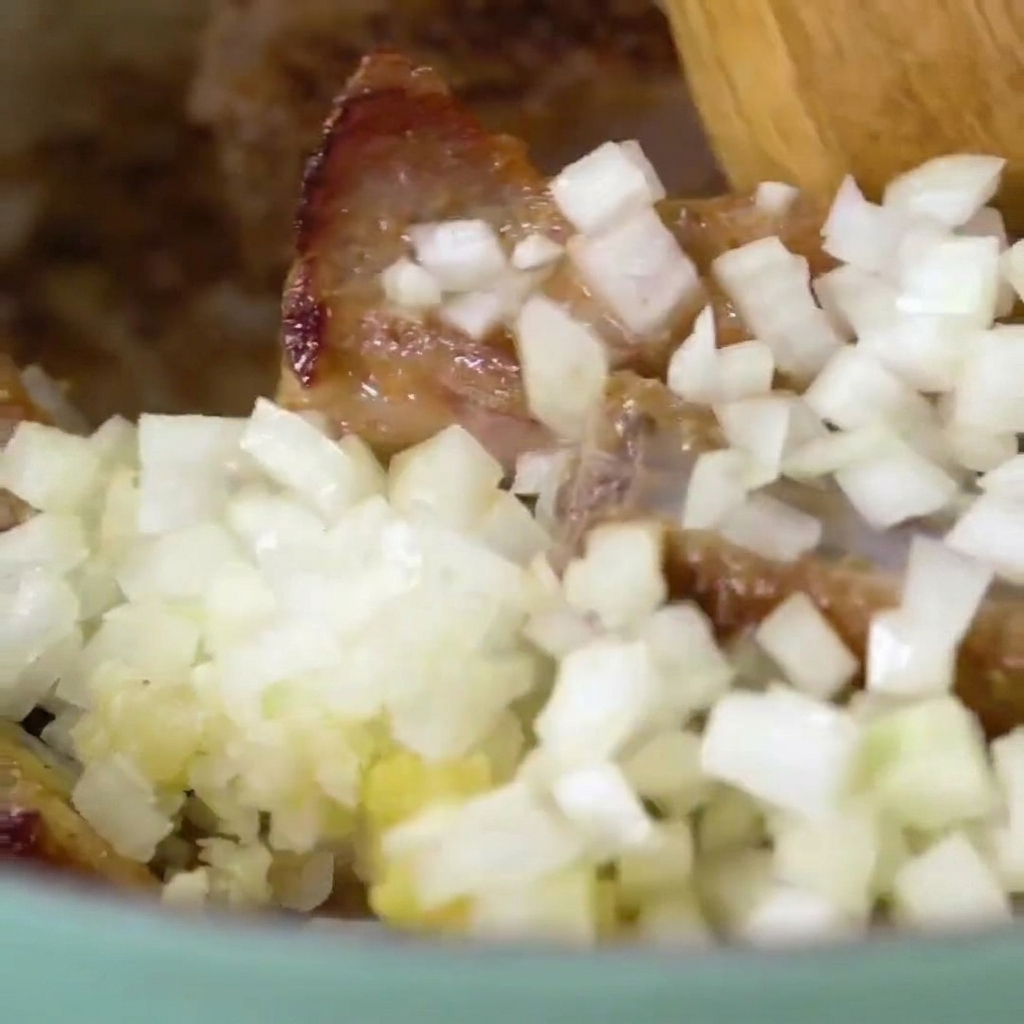 Nahaufnahme eines Holzlöffels, der gebräunte Rippchen mit Zwiebeln und Knoblauch verrührt.