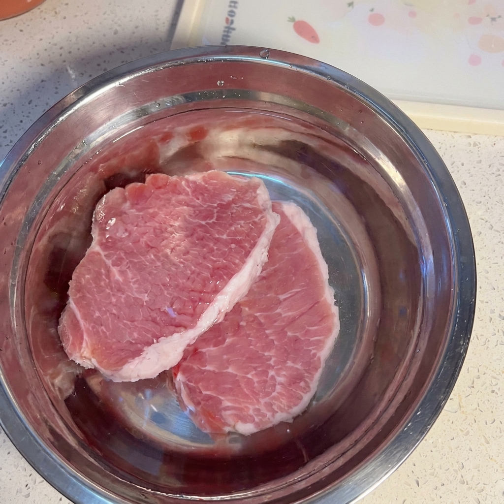 Rohe Schweinefleischscheiben in einer Metallschüssel mit Wasser.