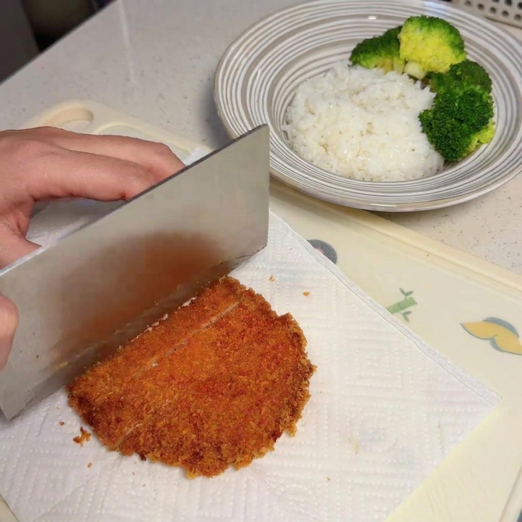 Eine Hand schneidet mit einem großen Messer ein knusprig goldbraun frittiertes Schweinekotelett auf Küchenpapier in Streifen.