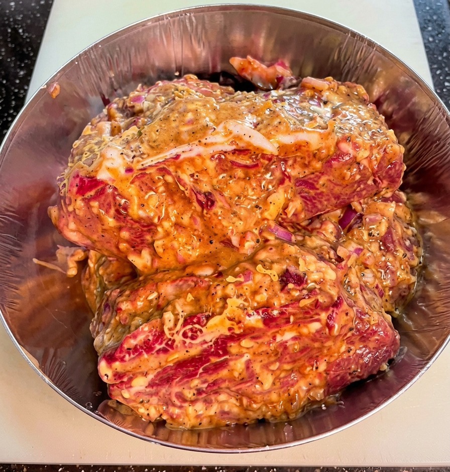 Nahaufnahme von rohen Fleischstücken in einer Metallschüssel, überzogen mit einer dicken Marinade mit sichtbaren Zwiebelstücken und grobem Pfeffer.