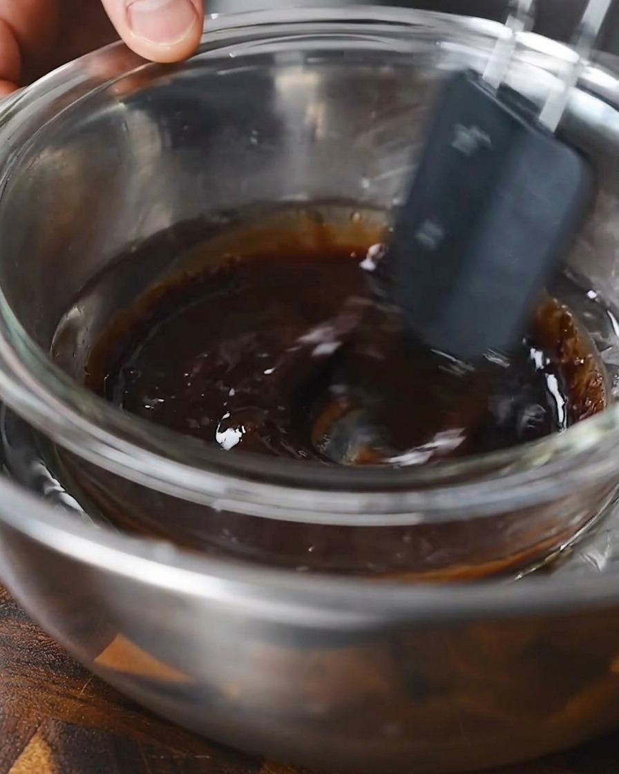Ein Spatel rührt eine dunkle flüssige Marinade in einer Glasschüssel über einem Wasserbad.