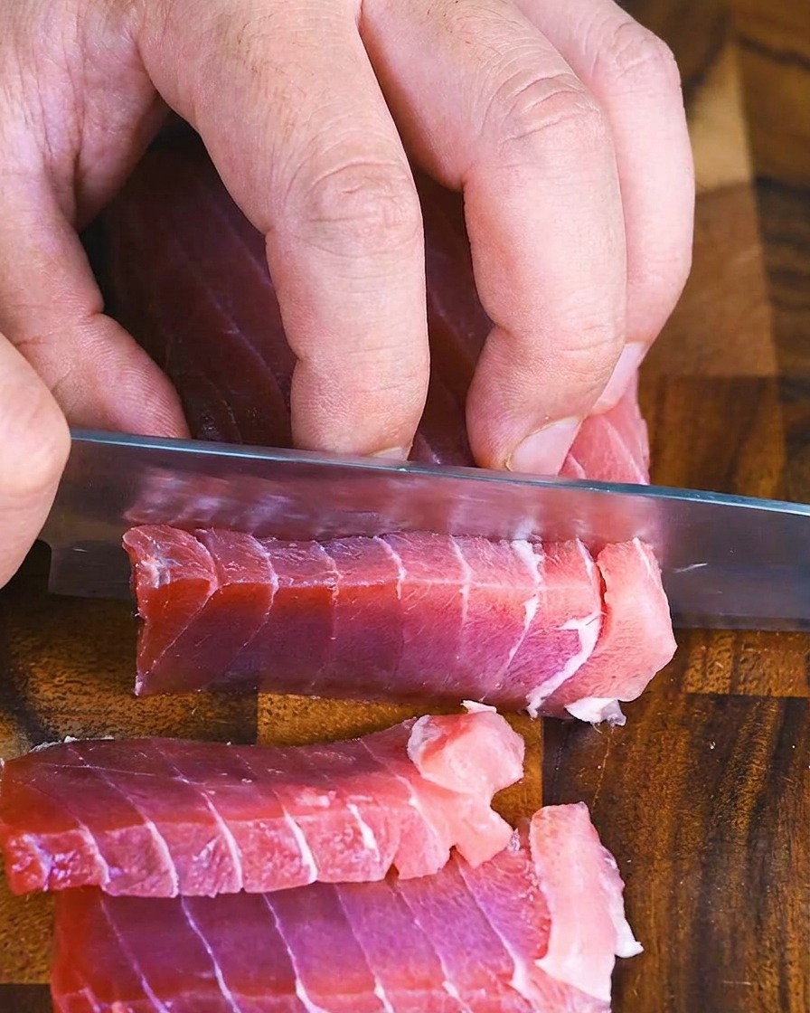 Hände verwenden ein Kochmesser, um einen Block rohen Thunfisch in dicke, gleichmäßige Stücke zu schneiden.