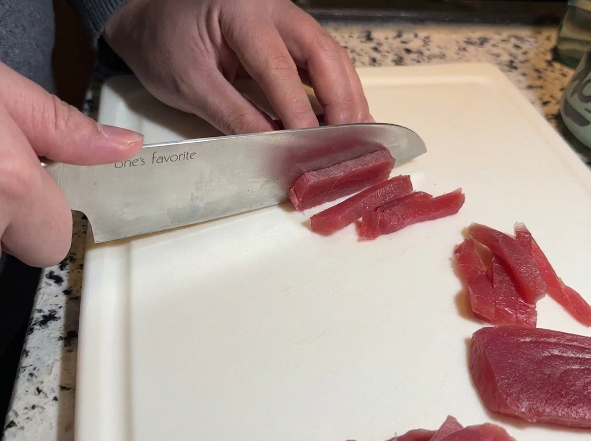 Ein scharfes Kochmesser schneidet leuchtend roten rohen Thunfisch auf einem weißen Schneidebrett in lange Streifen.
