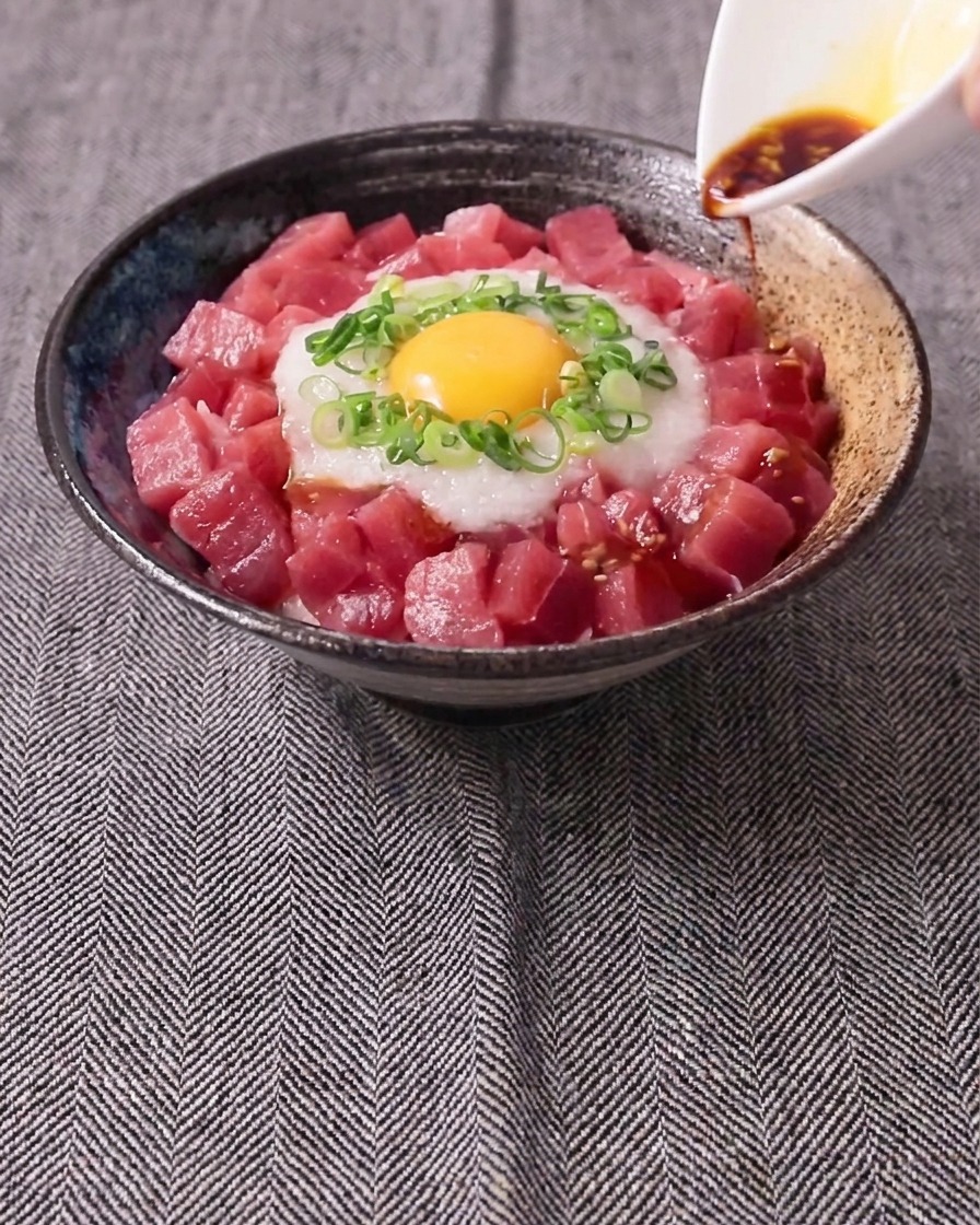 Dunkle Sauce aus einem kleinen weißen Kännchen über eine Schale mit Thunfisch, geriebener Yamswurzel, Frühlingszwiebeln und Eigelb.