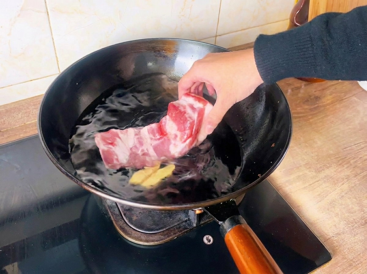 Eine Hand legt ein rohes Schweinebauchstück in einen mit kaltem Wasser und Ingwerscheiben gefüllten Wok.