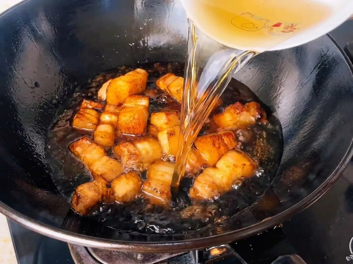 Aufgebrühtes Grünteewasser wird in einen Wok voller gewürzter Schweinebauchwürfel gegossen.