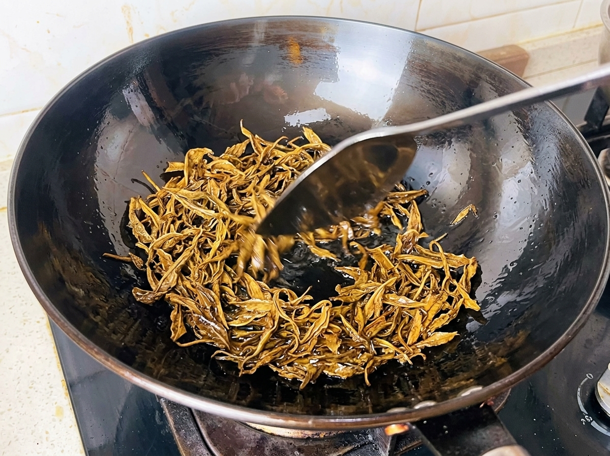 Feuchte Grünteeblätter werden in heißem Öl in einem schwarzen Wok mit einem Metallspatel frittiert.