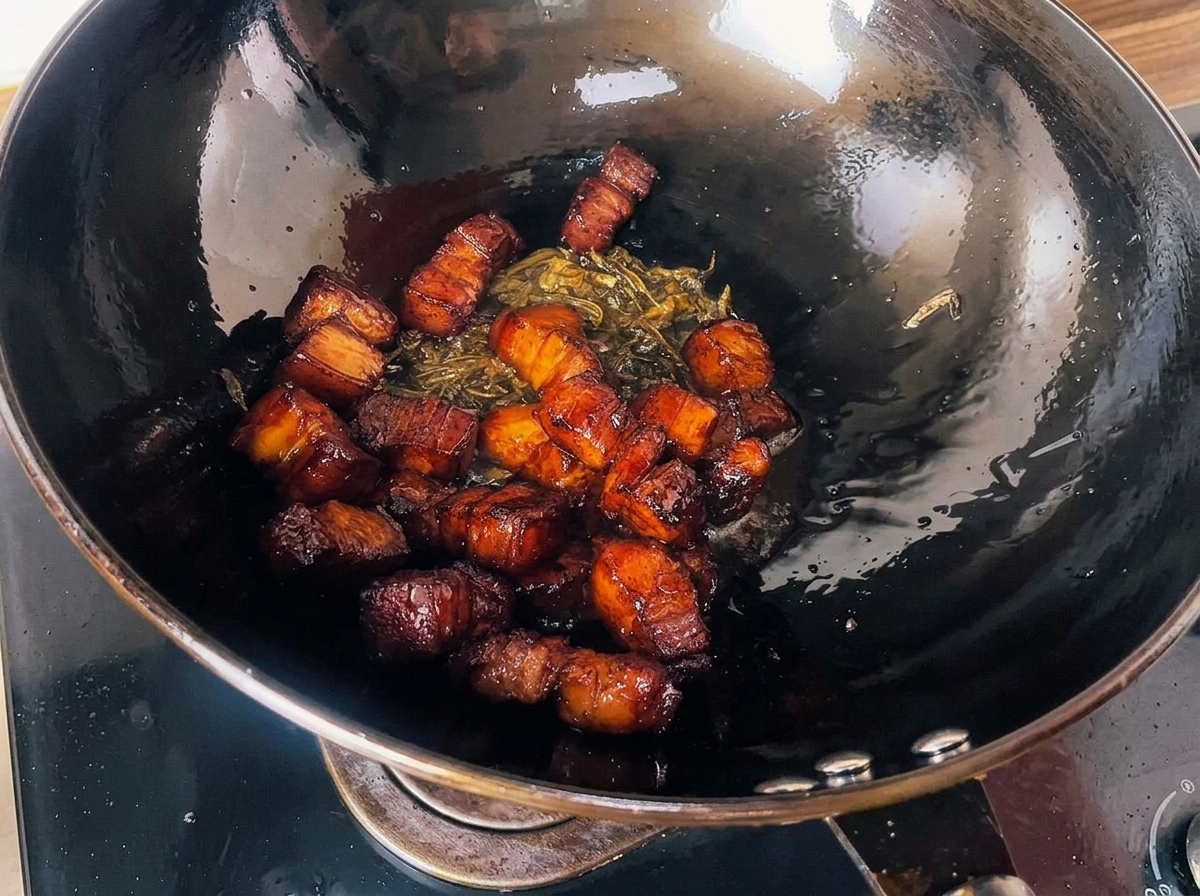 Geröstete, dunkel glasierte Schweinebauchwürfel vermischt mit knusprig gebratenen Teeblättern in einem großen schwarzen Wok.