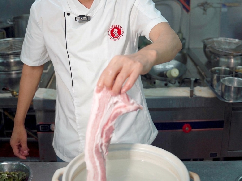 Ein Koch hält ein großes Stück rohen Schweinebauch über einen weißen Topf mit Wasser.