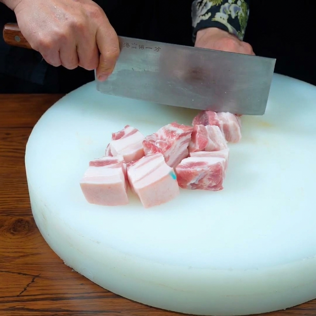 Hände schneiden mit einem Hackbeil einen Schweinebauch in gleichmäßige Würfel auf einem weißen Schneidebrett.