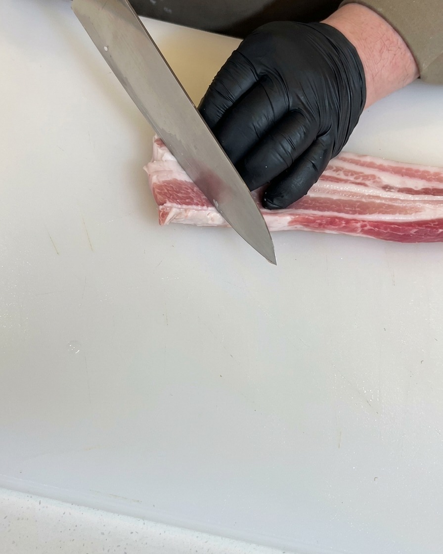 Hände mit schwarzen Handschuhen nutzen ein Kochmesser, um tiefe diagonale Schnitte in ein Stück rohen Schweinebauch auf einem weißen Schneidebrett zu machen.
