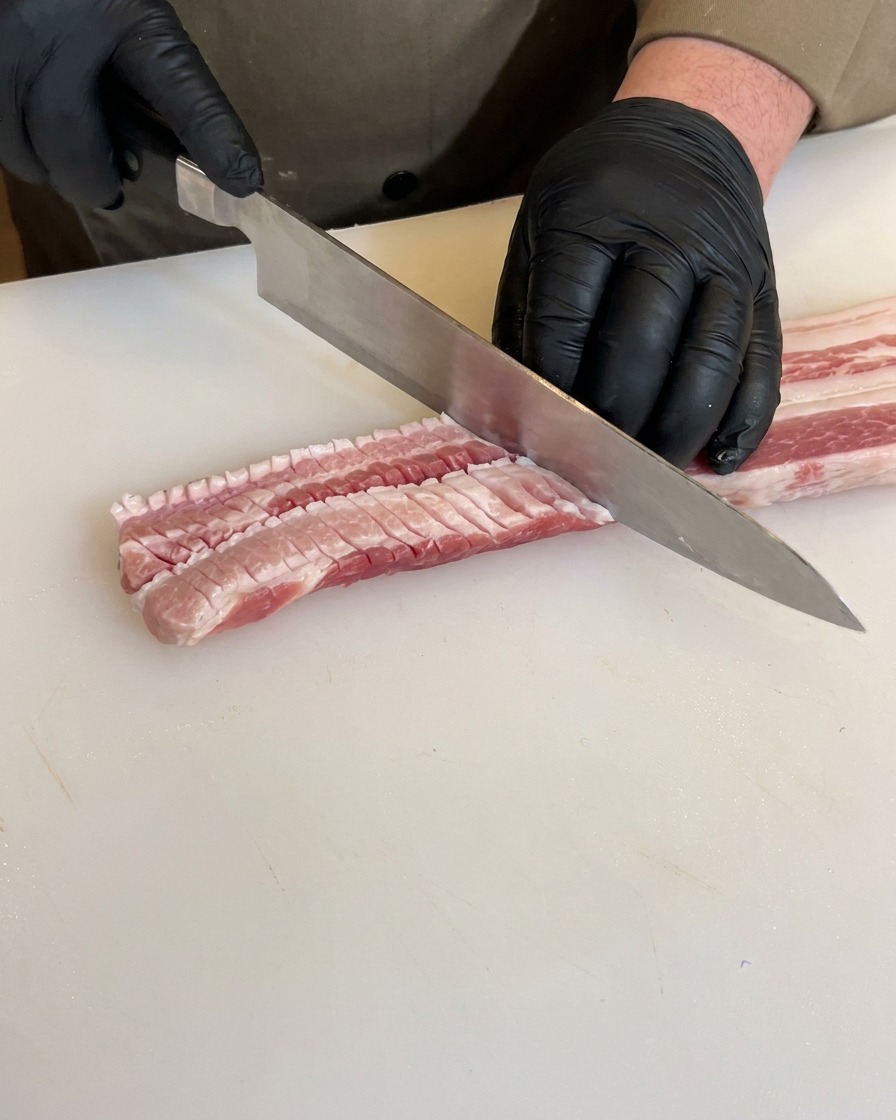 Hände in schwarzen Handschuhen schneiden die Rückseite eines rohen Schweinebauchs mit einem Kochmesser ein und zeigen tiefe, gitterartige Schnitte im Fleisch.