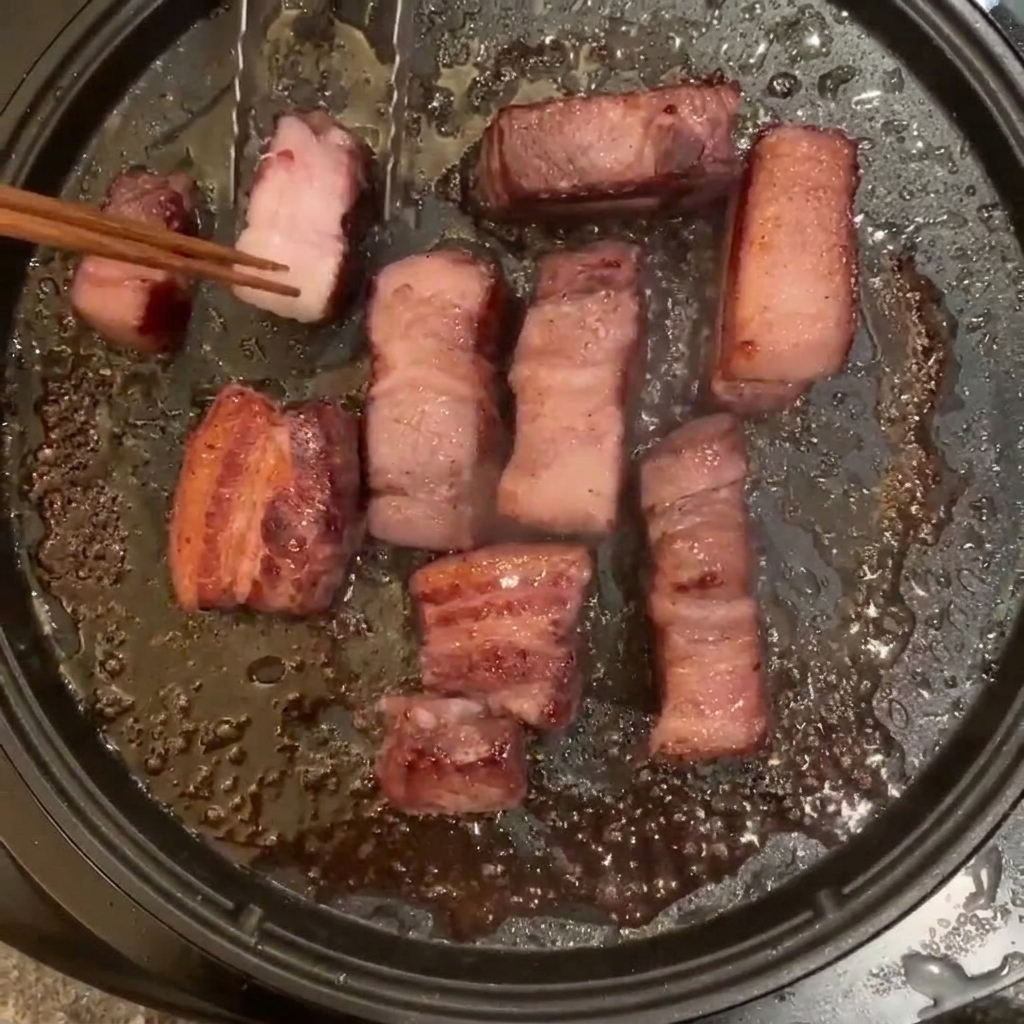 Schweinebauchstücke braten in einer Pfanne und werden mit Stäbchen gewendet.