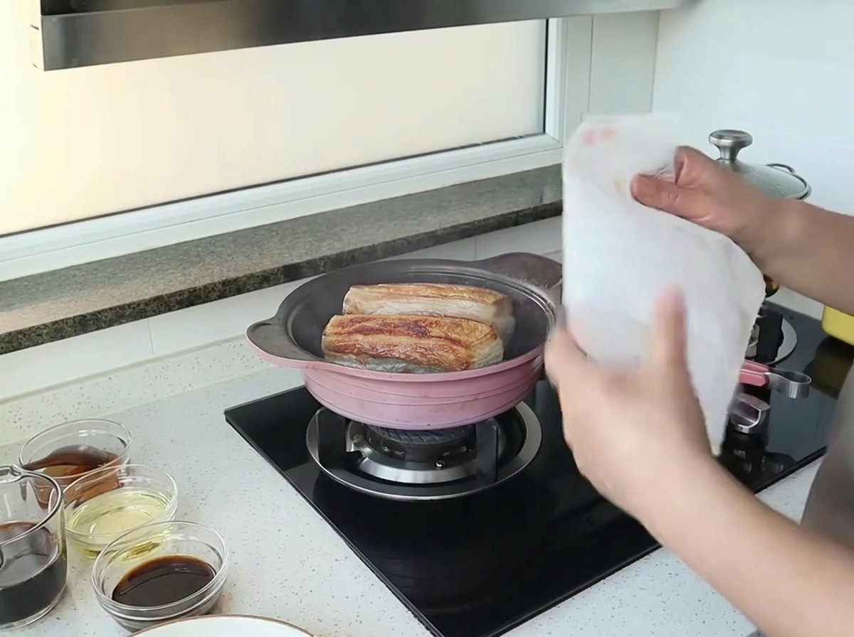 Hände halten ein weißes Küchenpapier über eine Pfanne mit zwei Stücken anbratendem Schweinebauch.