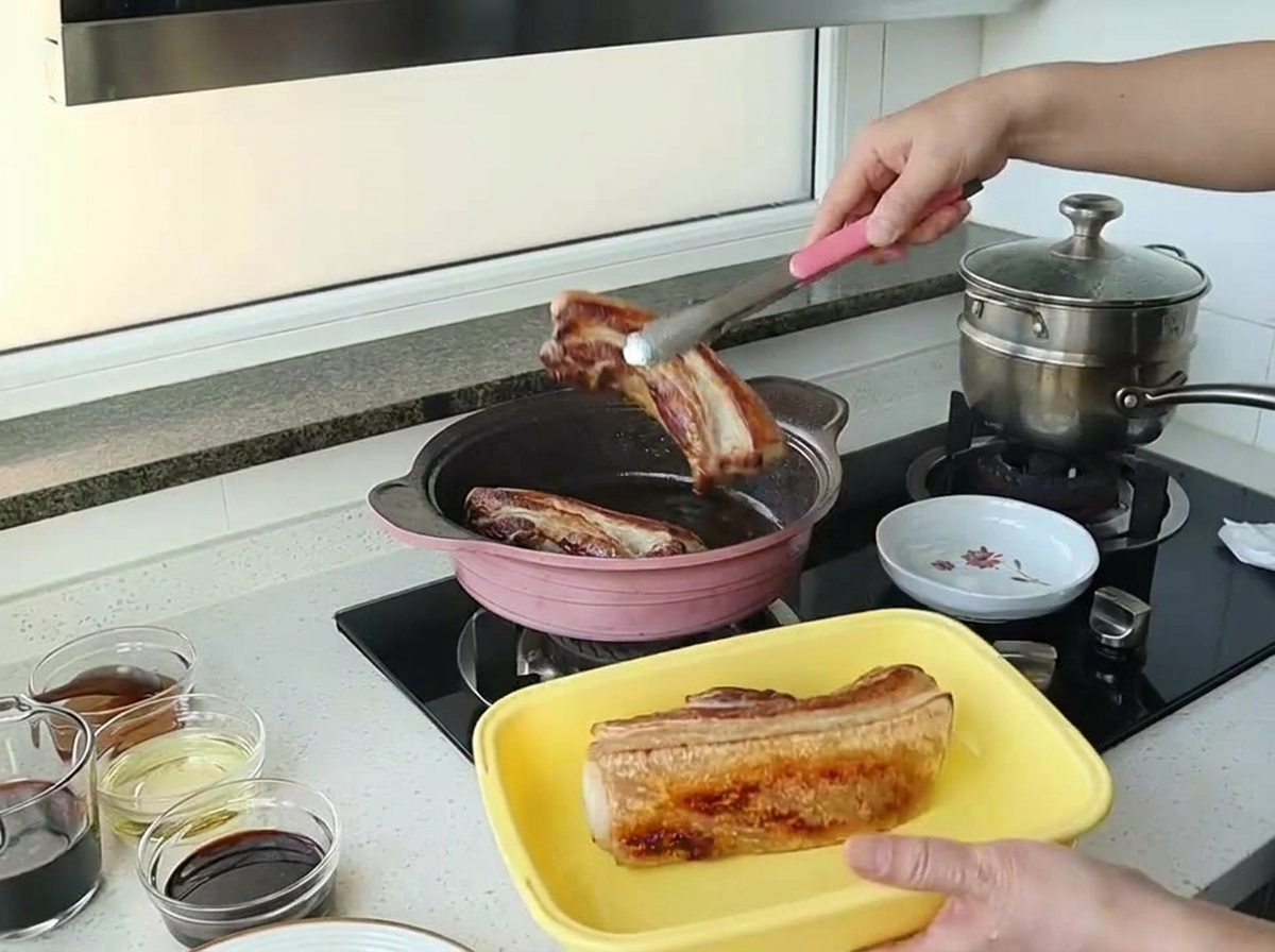 Eine Zange hebt ein wunderschön gebräuntes Stück Schweinebauch aus einer rosafarbenen Pfanne.