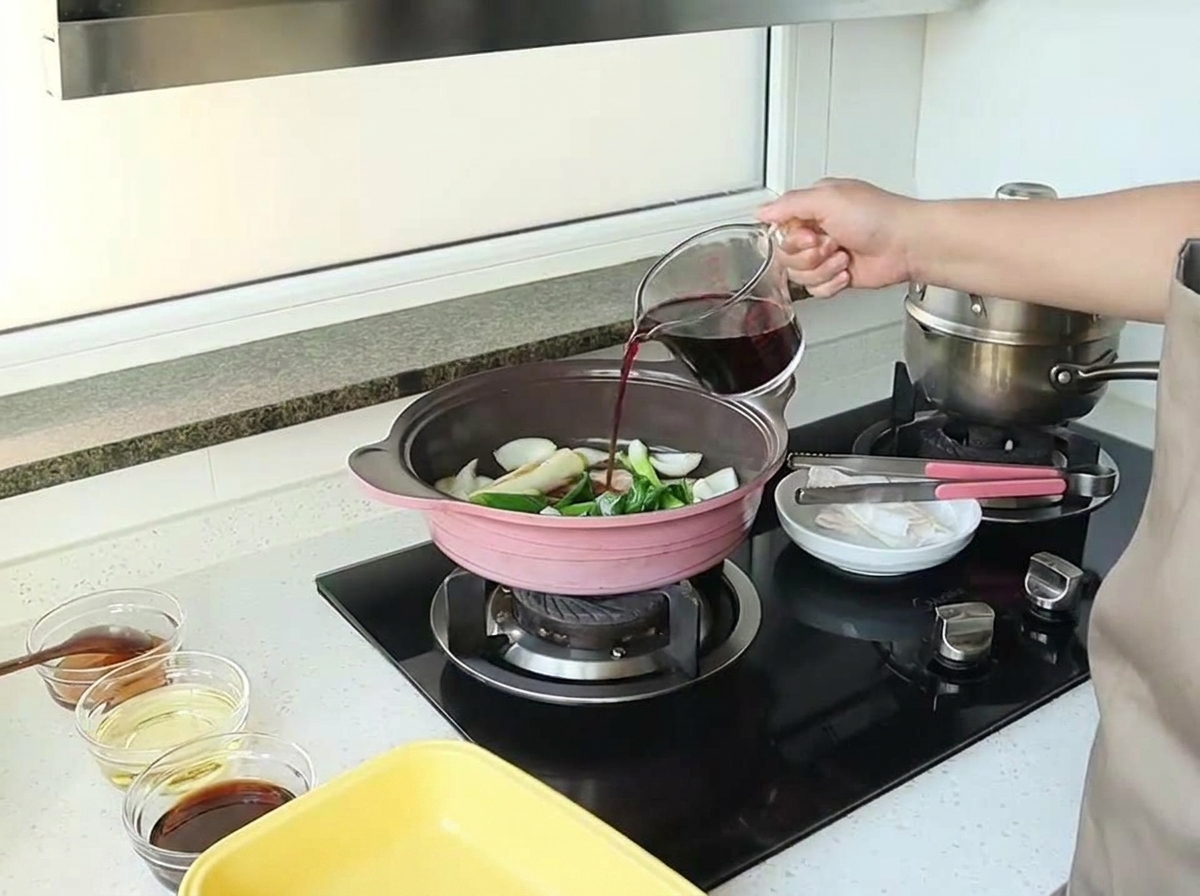 Eine Hand gießt Rotwein aus einem gläsernen Messbecher in eine Pfanne mit angeschwitzten Zwiebeln und Aromaten.