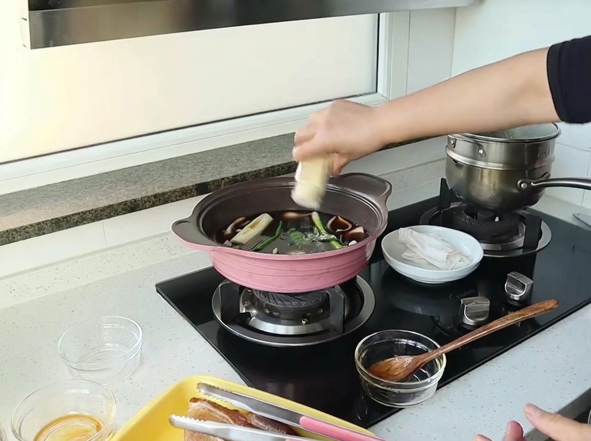 Eine Hand schüttelt Ingwerpulver aus einem kleinen Glas in einen Topf mit dunkler Sauce, Zwiebeln und Frühlingszwiebeln.