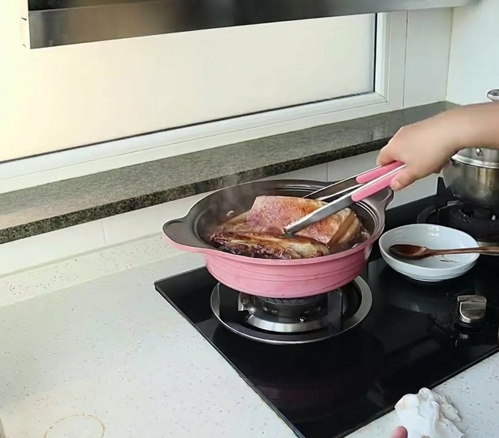Eine Person wendet mit einer Zange große Stücke Schweinebauch, die in einer dunklen Sauce in einem rosa Topf köcheln.