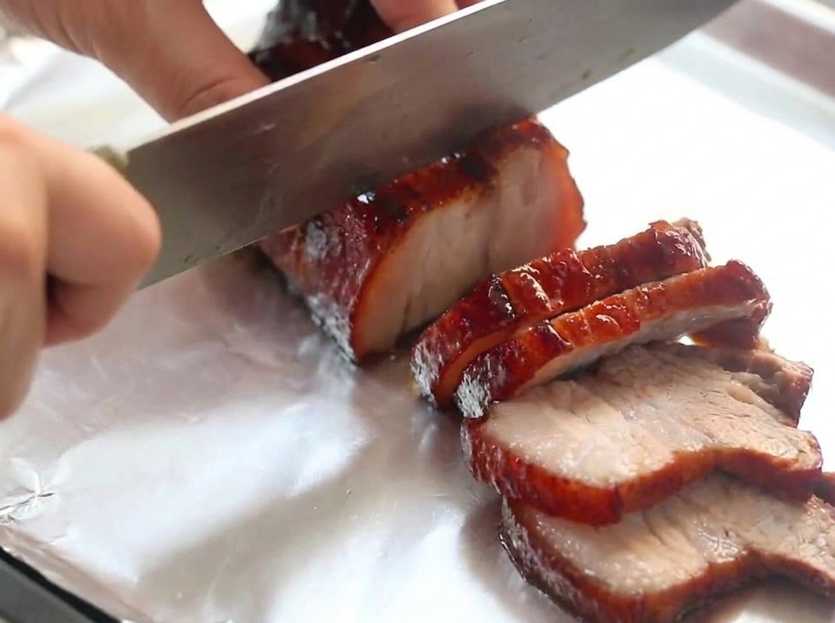 Ein Kochmesser schneidet durch ein Stück dunklen, in Rotwein glasierten Schweinebauch, das auf einem Stück Aluminiumfolie ruht.