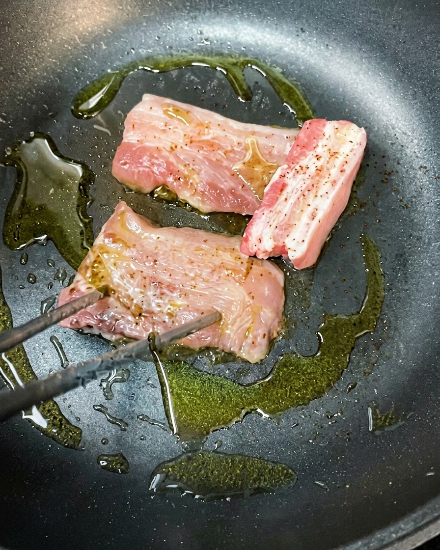 Marinierte Schweinebauchscheiben, die in einer heißen Pfanne mit Olivenöl gebräunt werdencite: 1.