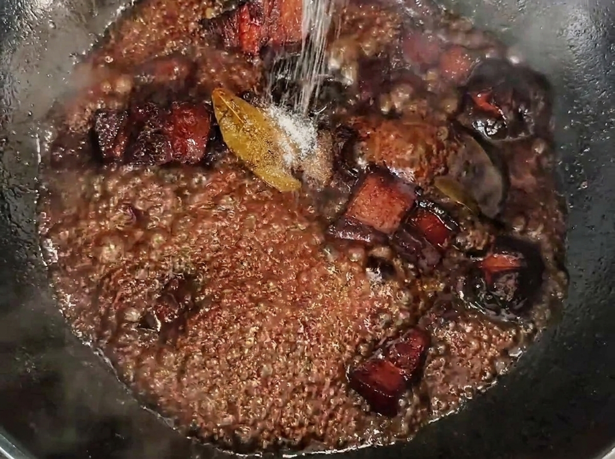 Salz wird über die schmorenden Schweinebauchwürfel in einer dunklen, reichhaltigen Sauce gestreut.