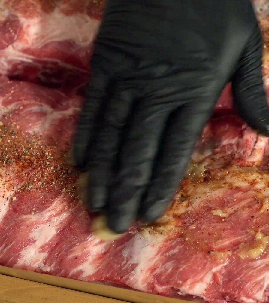 Eine Hand mit schwarzem Handschuh massiert Gewürze und Knoblauch in die rohen Schweinerippchen ein.