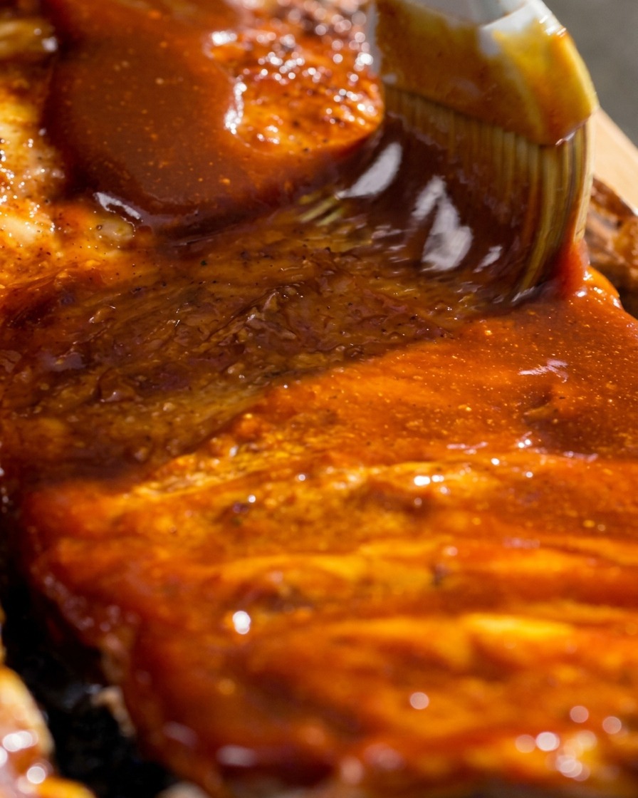 Nahaufnahme eines Pinsels, der dicke, glänzende rote Barbecue-Glasur auf die gegarten Rippchen streicht.