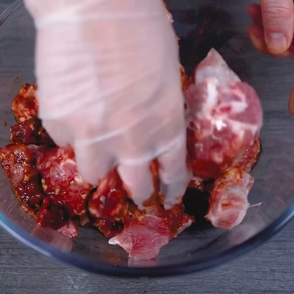 Eine behandschuhte Hand knetet die rote Marinade gründlich in die rohen Schweinerippchen in einer Glasschüssel.