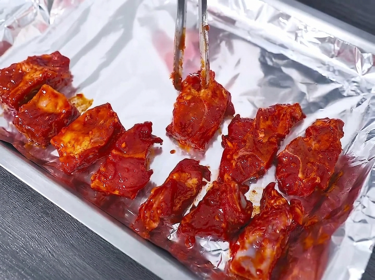 Rohe marinierte Schweinerippchen werden mit einer Metallzange ordentlich auf einem mit Alufolie ausgelegten Backblech angeordnet.