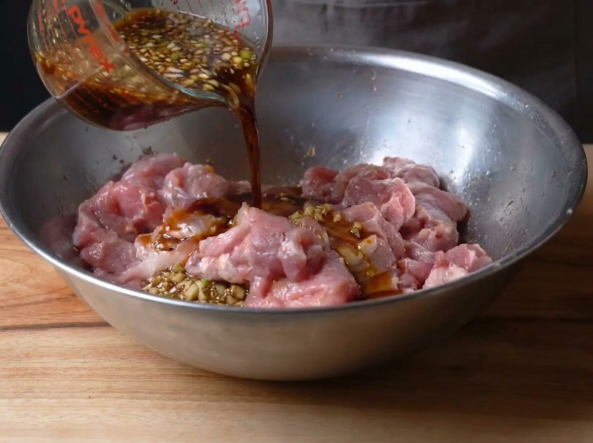 Die dunkle, herzhafte Marinade wird über das vormarinierte Schweinefleisch in einer Metallschüssel gegossen.