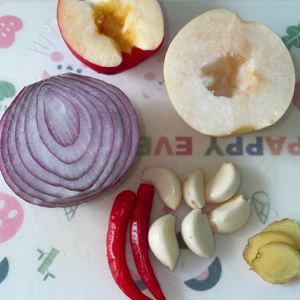 Halbierter Apfel, halbe rote Zwiebel, halbe Birne, Knoblauchzehen, rote Chilis und Ingwerscheiben auf einem Schneidebrett.
