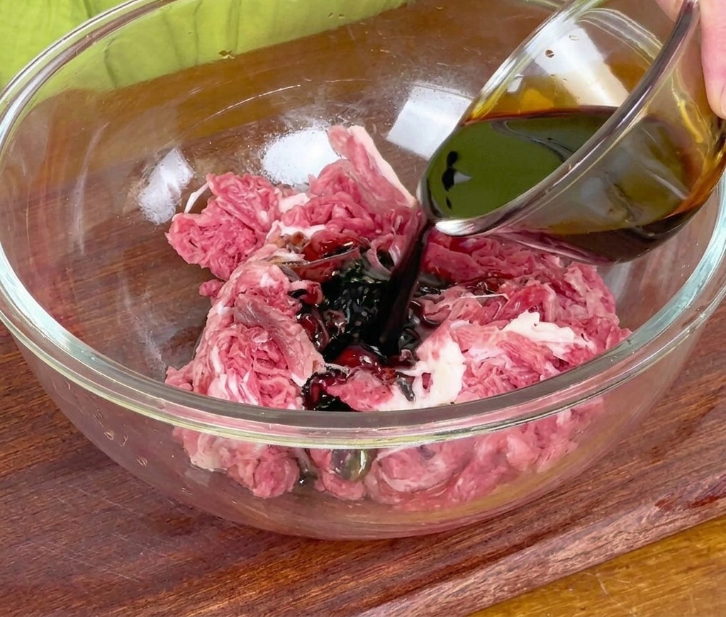 Dunkle Sojasauce wird aus einer kleinen Glasschale über rohes, in Scheiben geschnittenes Rindfleisch in einer großen Glasschüssel gegossen.