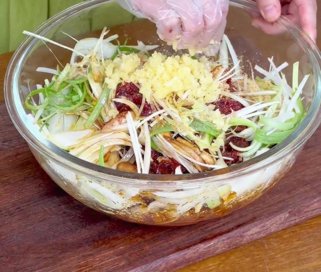 Eine Hand streut frisch gehackten Knoblauch über das Rindfleisch und die geschnittenen Zwiebeln in einer Glasschüssel.