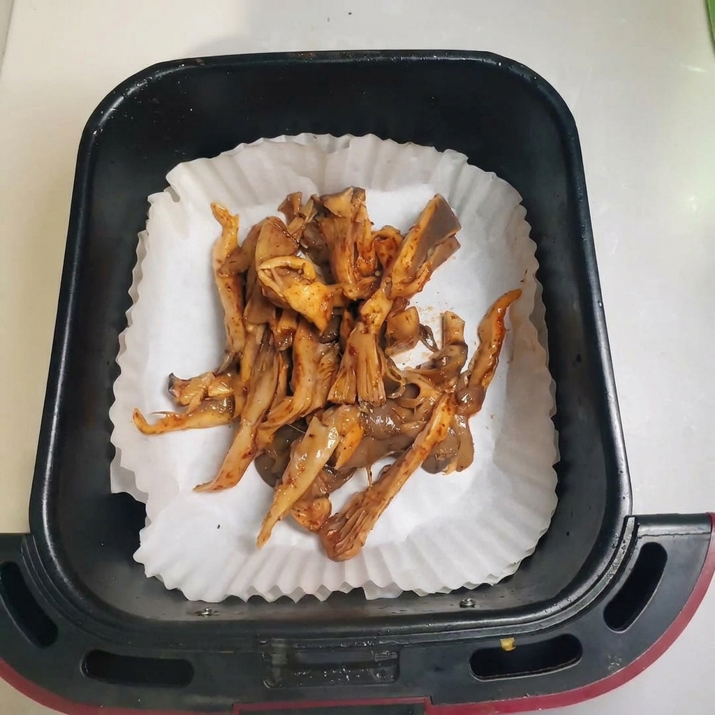 Gewürzte Pilze liegen verteilt in einem mit Backpapier ausgelegten Heißluftfritteusen-Korb.