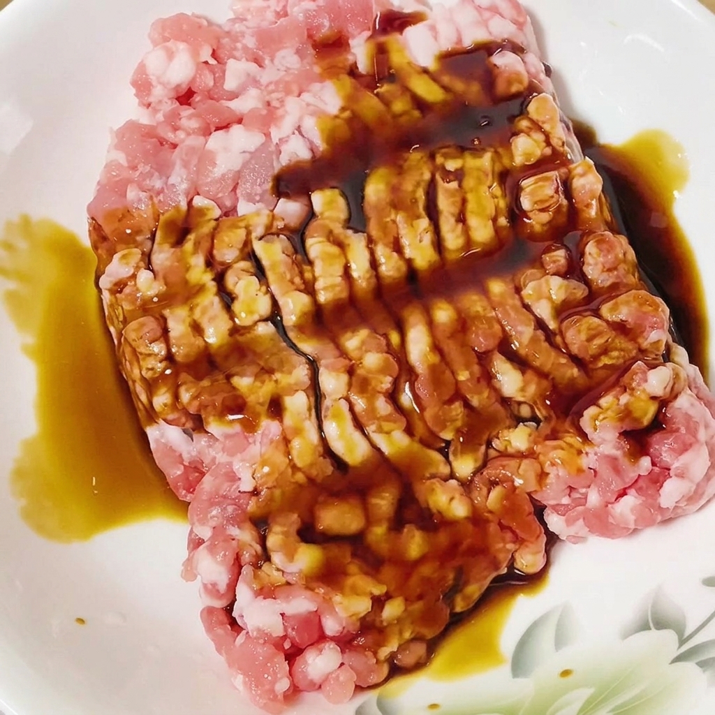 Sojasauce wird in einer weißen Schüssel über das rohe Schweinehackfleisch gegossen.
