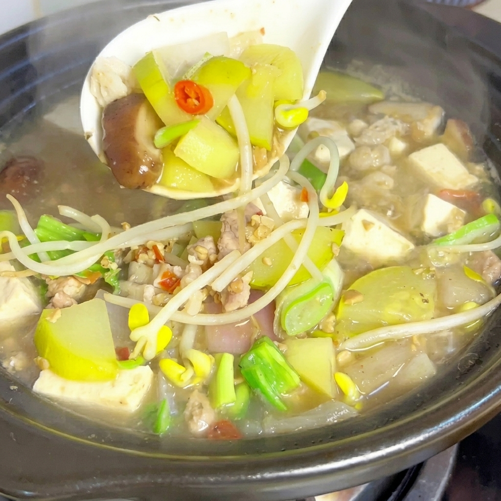 Eine weiße Schöpfkelle hebt vorsichtig gemischtes Gemüse, Tofu und Fleisch aus einem dampfenden, blubbernden Tontopf mit Sojabohnenpasten-Eintopf.