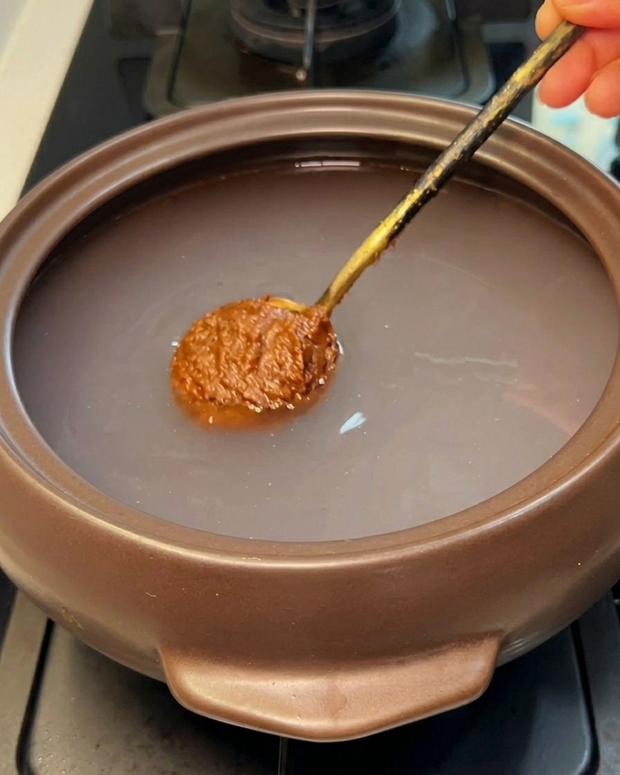 Ein Löffel brauner koreanischer Sojabohnenpaste wird in das klare Wasser eines Steintopfs getaucht.