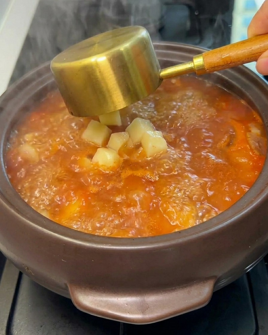 Rohe Kartoffelwürfel werden von einer Kelle in den kochenden Eintopf gegeben.