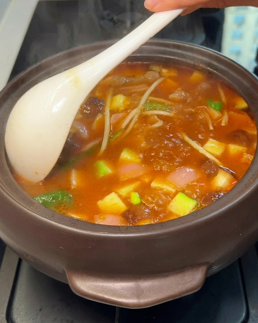Eine Nahaufnahme eines stark kochenden koreanischen Eintopfs in einem dunklen Topf, der mit einer weißen Kelle umgerührt wird, um Tofu, Zucchini, Pilze und Fleisch in einer orangeroten Brühe zum Vorschein zu bringen.