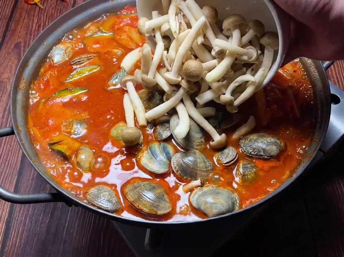 Frische Muscheln und verschiedene Pilzsorten werden in einen sprudelnden roten Kimchi-Eintopf gegeben.