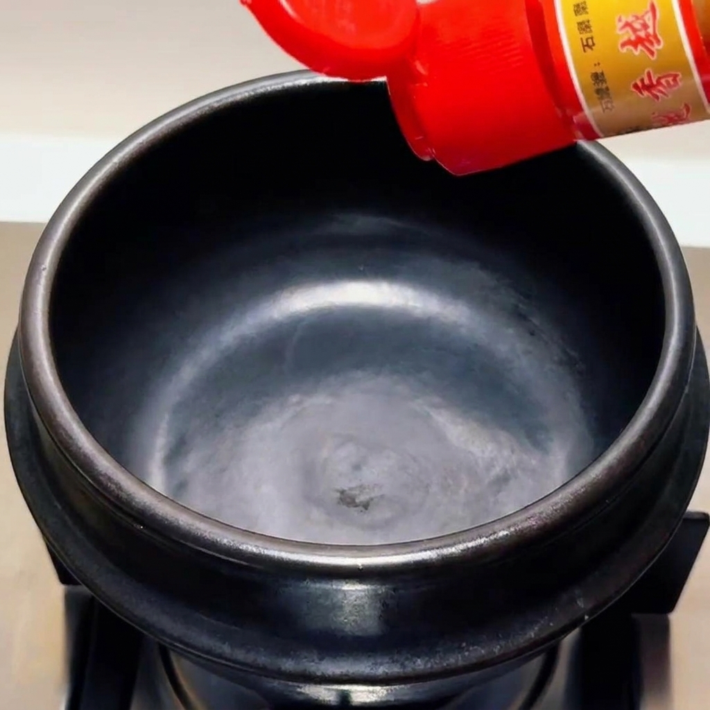 Sesamöl wird aus einer Flasche mit rotem Verschluss in einen leeren schwarzen Tontopf gegossen.