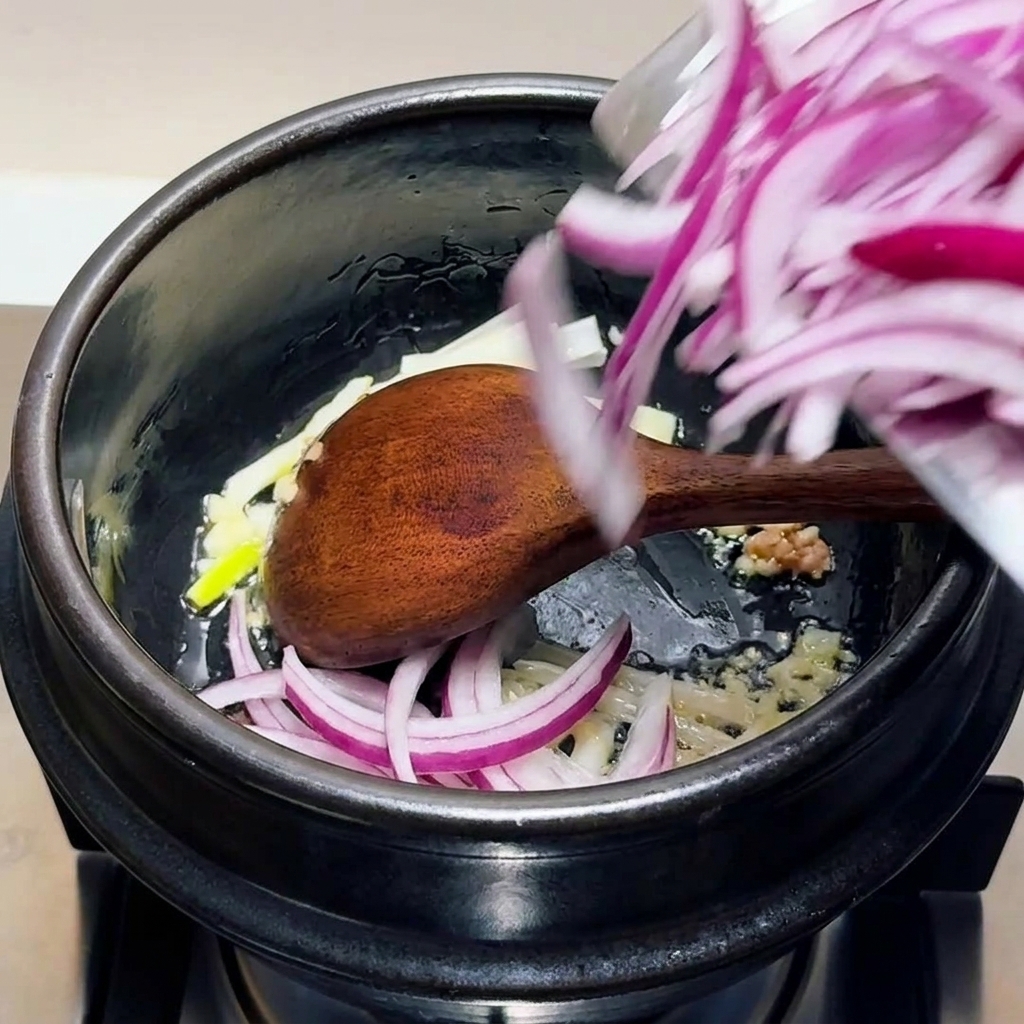 Geschnittene rote Zwiebeln, gehackter Knoblauch und Frühlingszwiebel-Weiß werden in einem schwarzen Topf mit einem Holzlöffel verrührt.
