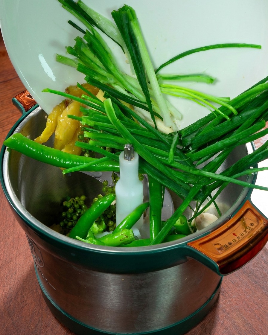 Ein Küchenmaschinenbehälter gefüllt mit Frühlingszwiebeln, Ingwer, Knoblauch, frischen grünen Chilis und Sichuan-Pfeffer.