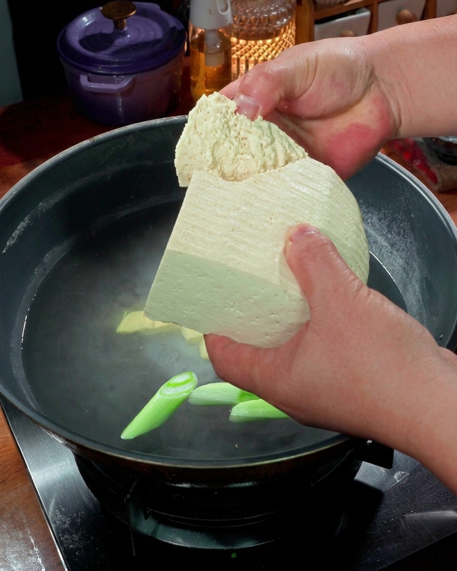 Hände, die große Stücke festen Tofu in einen Topf mit siedendem Wasser und Frühlingszwiebeln geben.