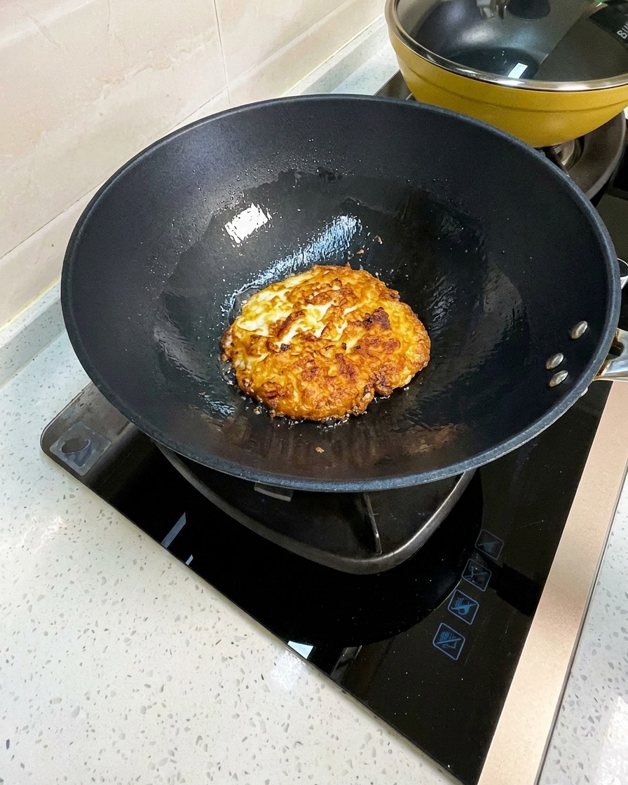 Die gebratenen Eier im Wok werden mit einem Metallpfannenwender in große Stücke zerteilt.