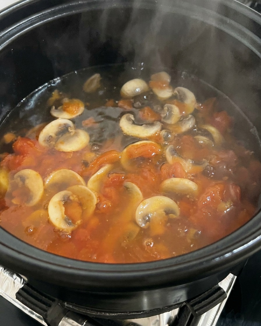 Kochendes Wasser wird in einen Topf mit angebratenen Tomaten und Pilzen gegossen, wobei Dampf entsteht.
